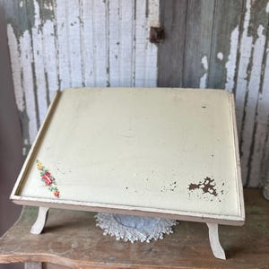 May include: An antique cream-colored lap desk with a floral design in the corner. The desk has a wooden frame and legs, and it sits on a wooden table. The desk is slightly angled, and the surface shows signs of wear.