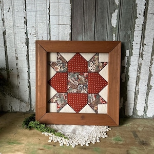 Peut inclure: Un bloc de courtepointe encadré, fait à la main, avec un motif d'étoile. L'étoile est composée de pièces de tissu rouge et à motifs floraux, sur un fond crème. Le cadre en bois carré a une finition marron chaud. Le tout est posé sur une surface en bois rustique.
