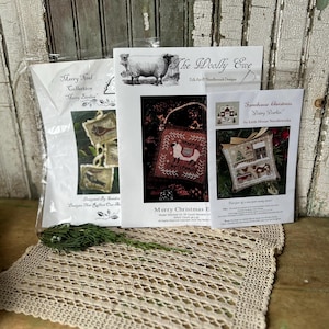 May include: Three cross-stitch pattern packets with Christmas themes, including "Merry Christmas E" and "Farmhouse Christmas." The packets are on a beige crocheted doily, with a wooden background.