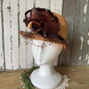 Può includere: Un cappello di paglia vintage con un ornamento marrone. Il cappello ha una corona rotonda e un'ampia tesa, con un velo di rete marrone scuro. Il cappello è esposto su una testa di manichino bianca.
