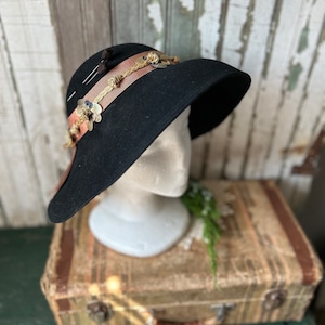 May include: A black felt cloche hat with a wide brim, decorated with a brown ribbon, gold accents, and delicate floral embellishments.