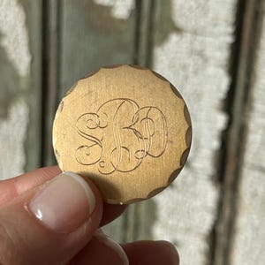 May include: A circular, gold-toned metal disc with a decorative, scalloped border. The disc displays an engraved monogram of the letters "SBD" in a flowing script. The background is a weathered wooden surface.