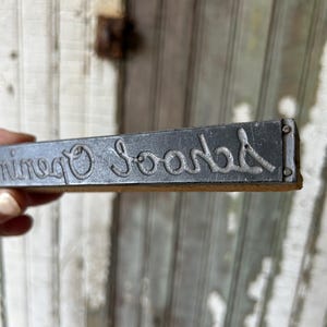 May include: A rectangular metal stamp with the word "Organic Soap" in a cursive font. The stamp is dark gray and appears to be vintage. The background is a weathered, light-colored wooden surface.