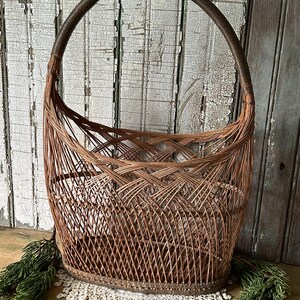 May include: A vintage, woven wicker basket with a large, arched handle. The basket is a warm brown color, with a detailed weave pattern. The handle is wrapped in a darker brown material. The basket sits on a white lace doily.
