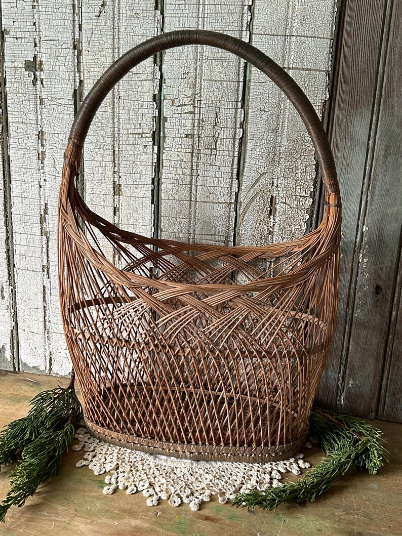 May include: A vintage, woven wicker basket with a large, arched handle. The basket is a warm brown color, with a detailed weave pattern. The handle is wrapped in a darker brown material. The basket sits on a white lace doily.