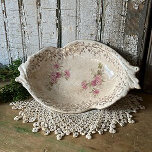 May include: An oval ceramic dish with a cream-colored base, featuring a delicate pink floral pattern in the center and intricate gold filigree detailing along the scalloped edges. The surface has a subtle crackle glaze finish throughout.