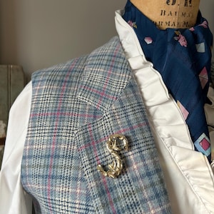 May include: A blue and white plaid vest with a gold-colored letter 'S' pin. The vest is worn over a white shirt with a ruffled collar and a blue patterned scarf.