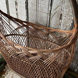 May include: A brown wicker basket with a curved handle. The basket has an oval base and a woven design. The background is a weathered, white-painted wooden surface. The basket is likely used for storage or display.