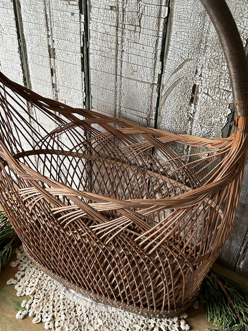 May include: A brown wicker basket with a curved handle. The basket has an oval base and a woven design. The background is a weathered, white-painted wooden surface. The basket is likely used for storage or display.