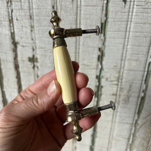 May include: Vintage cabinet pull with a cream-colored ceramic handle and brass-toned metal accents. The pull features decorative finials and includes screws for installation. A classic hardware piece for furniture restoration or new projects.
