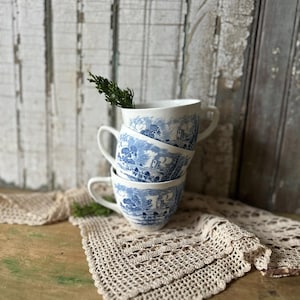 May include: A stack of three white ceramic teacups featuring a blue transferware pattern depicting a landscape with buildings and trees. A small green sprig is tucked into the top cup, and the stack rests on a beige crocheted lace doily.