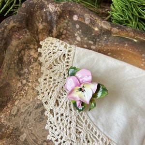 May include: A delicate porcelain flower brooch with a pink and white pansy design. The brooch is set on a white fabric with a crocheted lace trim.