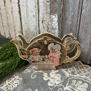 May include: A decorative teapot-shaped ornament with a gold trim and a vintage illustration of two children at a tea party. The ornament is set against a rustic wooden background and rests on a lace surface.