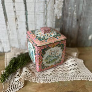 May include: A vintage pink metal box with a square shape and a matching lid. The box features a decorative floral and landscape design in shades of pink, green, and blue. The box is placed on a cream-colored lace doily.