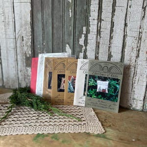 Op de afbeelding: Een verzameling 'Christmas of Olde'-boekjes met diverse omslagontwerpen, waaronder afbeeldingen van een huis, kerstbomen en groen. De boekjes liggen op een houten oppervlak met een gehaakte placemat en een takje groen.