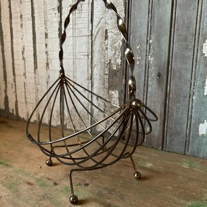 May include: A vintage metal wire basket with a twisted handle and a fan-like design. The basket has three small ball feet and a bronze finish. The basket is likely used for decorative purposes or to hold small items.
