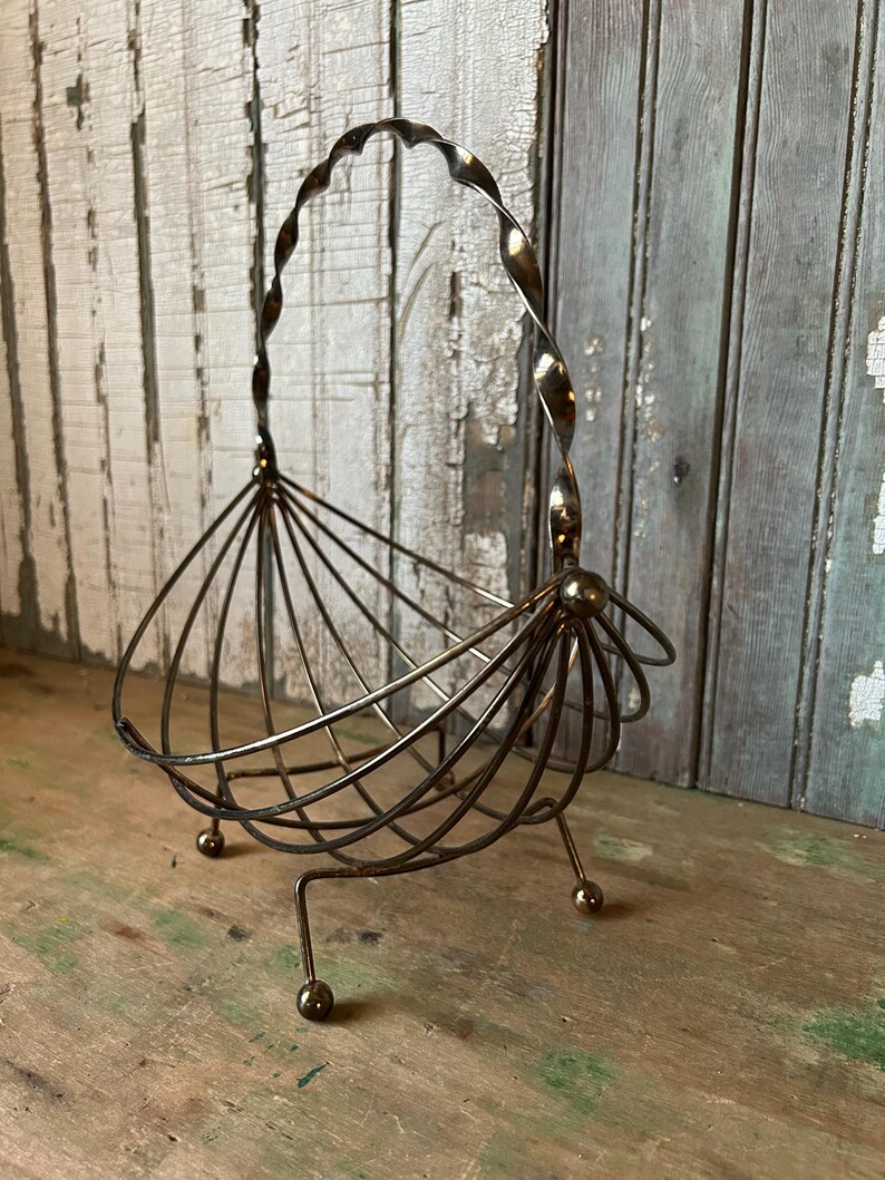May include: A vintage metal wire basket with a twisted handle and a fan-like design. The basket has three small ball feet and a bronze finish. The basket is likely used for decorative purposes or to hold small items.