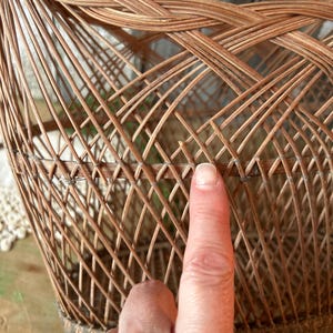 May include: A woven wicker basket with a rounded shape. The basket is made of thin, light brown wicker strips, intricately woven to create a lattice pattern. The base of the basket is reinforced with a darker, thicker material. The basket is empty.