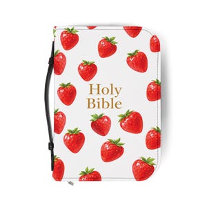 May include: A white Bible cover with a pattern of red strawberries and green leaves. The words "Holy Bible" are printed in gold. A black strap is attached to the side.