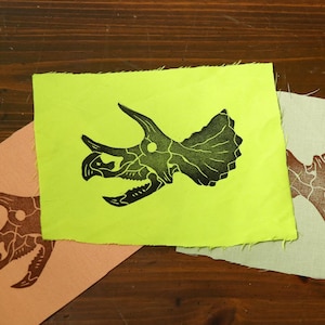 May include: Three rectangular fabric patches featuring a black triceratops skull print. One patch is neon yellow, one is light peach, and one is pale green. Each patch has raw, unfinished edges and displays a detailed dinosaur fossil graphic.