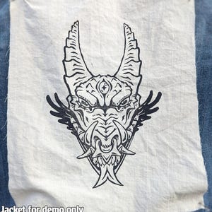 May include: A denim jacket with a white patch showing a black illustration of a dragon's head with horns and a third eye. The dragon has wing-like features. The text "Jacket for demo only" is at the bottom.