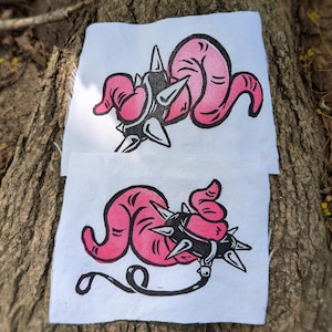 May include: Two white fabric squares with a pink and black linocut print. The print features a stylized pink worm-like creature with a spiked ball and a black line. The squares are placed on a tree trunk.