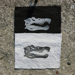 May include: Two rectangular pieces of fabric, one black and one white, each featuring a stylized alligator skull print in contrasting colors. The alligator skull is detailed with sharp teeth and eye sockets. The fabric pieces are placed side by side.