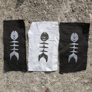 Peut inclure: Trois patchs en tissu avec un motif d'arête de poisson. Deux sont noirs avec une arête de poisson blanche, et un est blanc avec une arête de poisson noire. Le motif d'arête de poisson comprend un visage souriant et une queue stylisée.