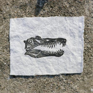 May include: A black and white linocut print of an alligator skull on a piece of white fabric. The alligator's jaws are open, revealing sharp teeth. The fabric has a raw edge and is set against a textured, light brown surface.