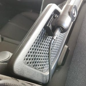 May include: A black dash camera mounted on a perforated black surface inside a vehicle. The camera is connected to a power source via a black cable. The camera lens is visible.