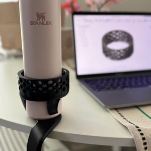May include: A light pink Stanley travel mug with a black cup holder attached to a black table clip. The mug has a silver rim and a dark pink lid. The Stanley logo is printed on the front of the mug. A laptop and flowers are in the background.