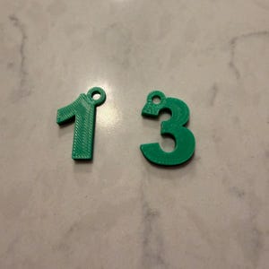 May include: Two green plastic charms shaped like the numbers one and three. Each number features a small circular loop at the top for attachment. The surface of the numbers displays a visible layered texture consistent with 3D printed objects.