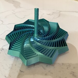 May include: A teal-colored, geometric 3D-printed top. The top has a star-like base with layered, angled edges and a central, ridged spindle. The design creates a spiral effect, showcasing a modern, artistic aesthetic.