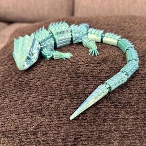 May include: A 3D printed articulated lizard figurine in a shimmering metallic light blue and teal color. The lizard features a segmented body with a textured, spiky scale pattern along its back and a long, flexible tail that curves to the side.