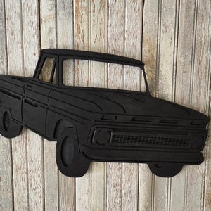Puede incluir: Recorte de madera negro de una camioneta pickup vintage. La camioneta presenta un diseño detallado con ruedas, cabina y caja de carga visibles. El recorte está montado sobre un fondo de tablones de madera.