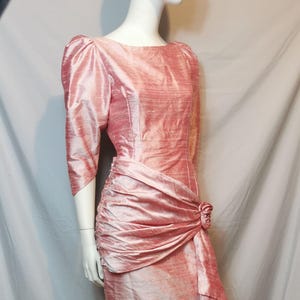 May include: A vintage, knee-length dress in a soft, dusty rose colour. The dress features a ruched waist detail, a draped side accent, and a decorative rose embellishment. It has three-quarter sleeves and a round neckline, displayed on a mannequin.