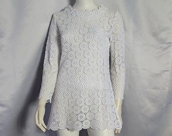 May include: A white long-sleeved mini dress featuring an all-over floral lace pattern with a scalloped neckline, cuffs, and hem. The garment has a high neckline and a straight silhouette, constructed from a semi-sheer white lace fabric.