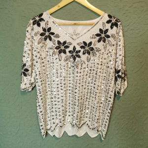 May include: Cream-coloured, short-sleeved top with a V-neck, decorated with black and silver sequins in a floral design. The top is hanging on a wooden hanger. The hemline has a scalloped edge. The background is green.