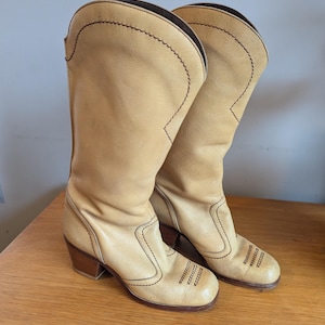 Op de afbeelding: Een paar lichtbruine leren cowboylaarzen met bruine stiksels en een blokhak. De laarzen hebben een ronde neus en een hoge schacht, met decoratieve stiksels op de voet en de schacht.