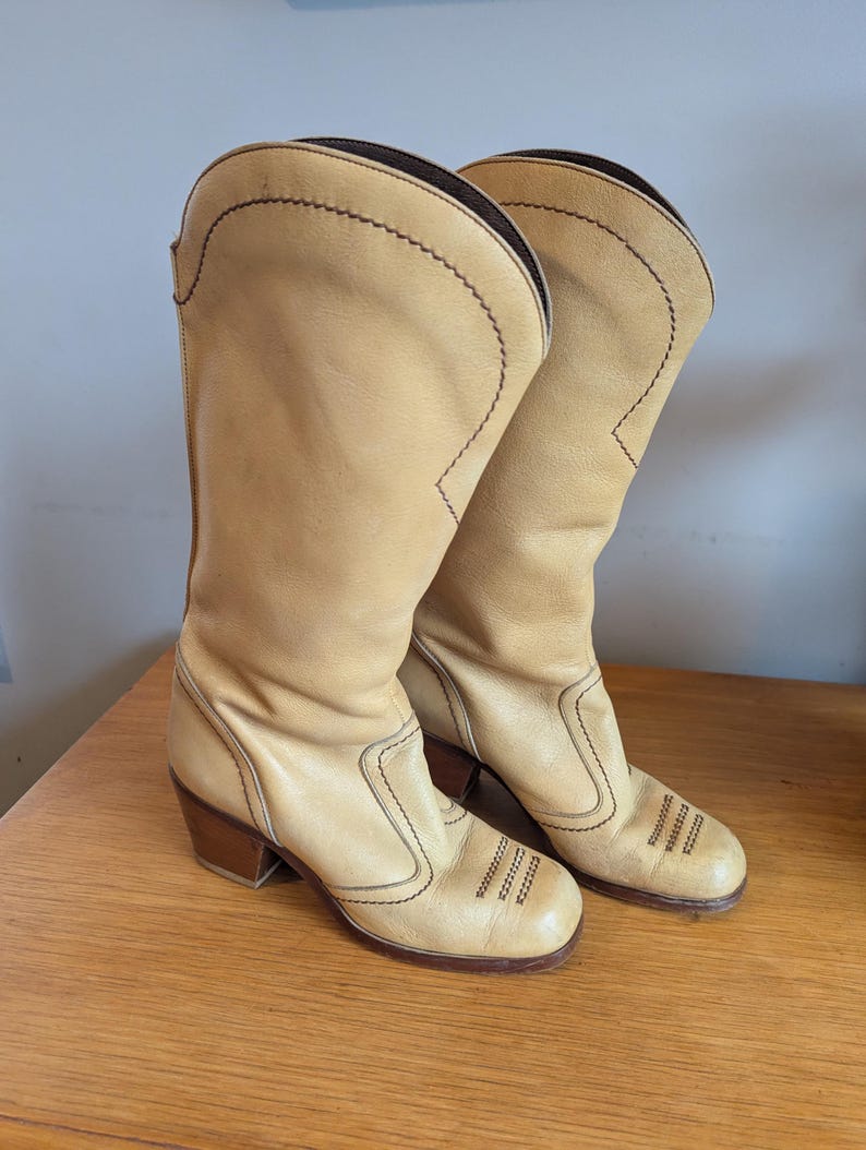Op de afbeelding: Een paar lichtbruine leren cowboylaarzen met bruine stiksels en een blokhak. De laarzen hebben een ronde neus en een hoge schacht, met decoratieve stiksels op de voet en de schacht.