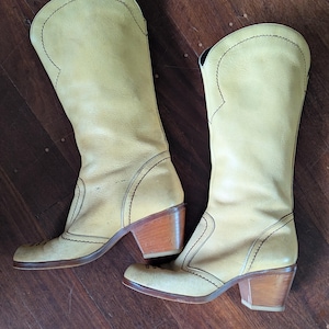 Op de afbeelding: Een paar vintage lichtgele leren cowboylaarzen. De laarzen hebben een hoge schacht, decoratieve stiksels en houten blokhakken. De zolen en hakken vertonen gebruikssporen. De laarzen staan op een houten oppervlak.