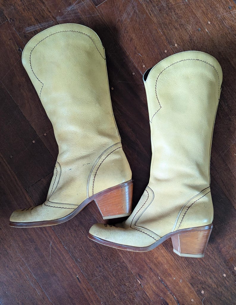 Op de afbeelding: Een paar vintage lichtgele leren cowboylaarzen. De laarzen hebben een hoge schacht, decoratieve stiksels en houten blokhakken. De zolen en hakken vertonen gebruikssporen. De laarzen staan op een houten oppervlak.