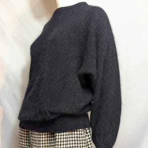 May include: A long-sleeved black sweater with a fuzzy, textured finish and a crew neckline. The sweater features ribbed cuffs and a ribbed hem, paired with a skirt that has a black and white houndstooth pattern visible at the bottom.