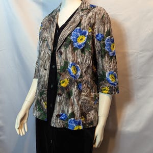 Può includere: Un blazer a maniche a tre quarti con un motivo marrone, grigio e verde simile alla corteccia. Il blazer presenta grandi accenti floreali blu e gialli. Ha un colletto a lancia, due tasche anteriori e bottoni marroni. Il blazer è indossato sopra un vestito nero.