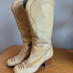 Op de afbeelding: Een paar vintage beige leren cowboylaarzen. De laarzen hebben een hoge schacht, decoratieve stiksels en een gestapelde houten hak. De laarzen worden op een houten oppervlak gepresenteerd.