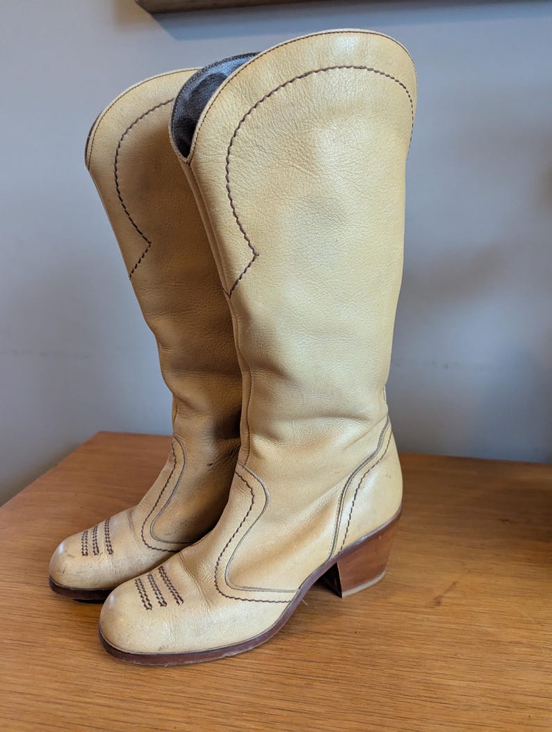 Op de afbeelding: Een paar vintage beige leren cowboylaarzen. De laarzen hebben een hoge schacht, decoratieve stiksels en een gestapelde houten hak. De laarzen worden op een houten oppervlak gepresenteerd.