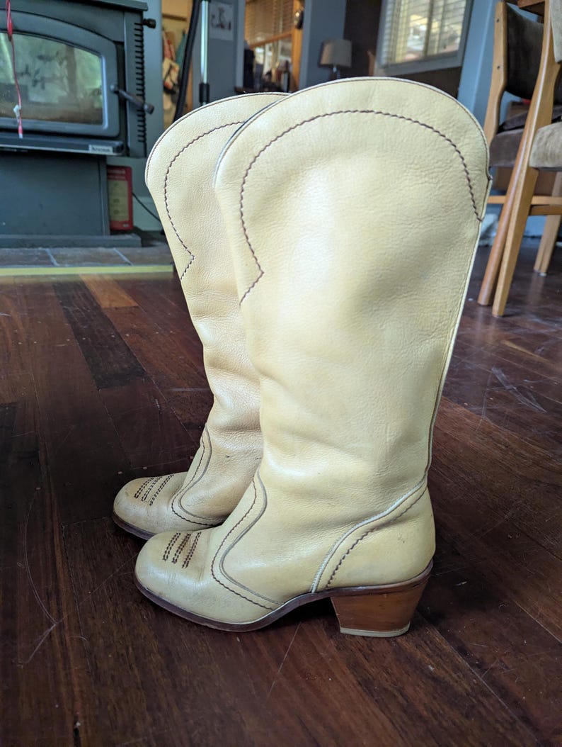 Op de afbeelding: Een paar cr&egrave;mekleurige vintage cowboylaarzen. De laarzen hebben een hoge schacht, decoratieve stiksels en een gestapelde houten hak. Het leer lijkt versleten, wat suggereert dat ze geliefd zijn en een klassieke Western-stijl hebben.
