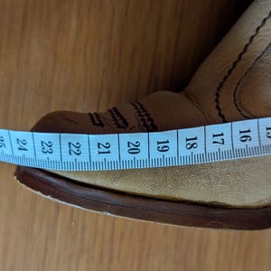 Op de afbeelding: Close-up van een bruinleren cowboylaars met decoratieve stiksels. Een blauwe meetlint toont een lengte van ongeveer 23 cm. De laars heeft een puntige neus en een gestapelde hak, wat duidt op een Western-thema.