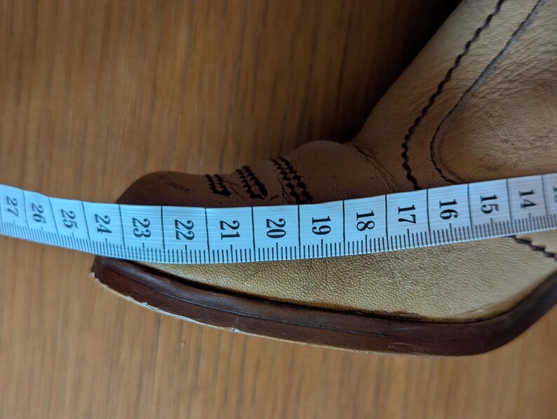 Op de afbeelding: Close-up van een bruinleren cowboylaars met decoratieve stiksels. Een blauwe meetlint toont een lengte van ongeveer 23 cm. De laars heeft een puntige neus en een gestapelde hak, wat duidt op een Western-thema.