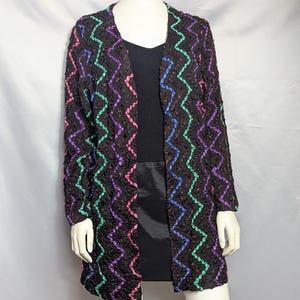 May include: A black open-front long-sleeve jacket featuring a textured surface and vertical zigzag patterns in alternating neon pink, bright green, and royal blue. The jacket is worn over a black ribbed top and a black skirt on a mannequin.